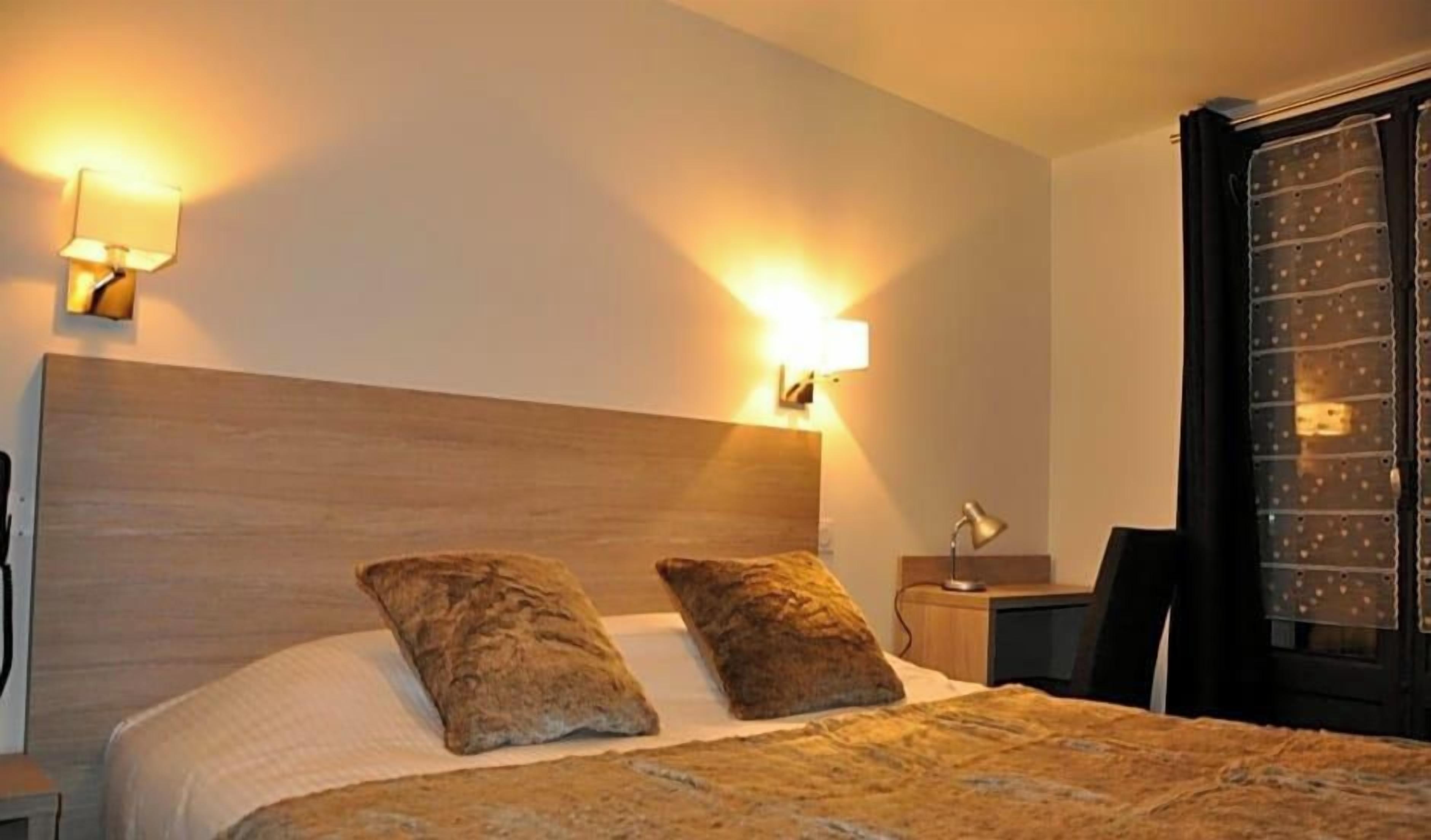 Une chambre d’hôtel avec une tête de lit en bois, deux oreillers, une lampe de chevet et une fenêtre avec un rideau.
