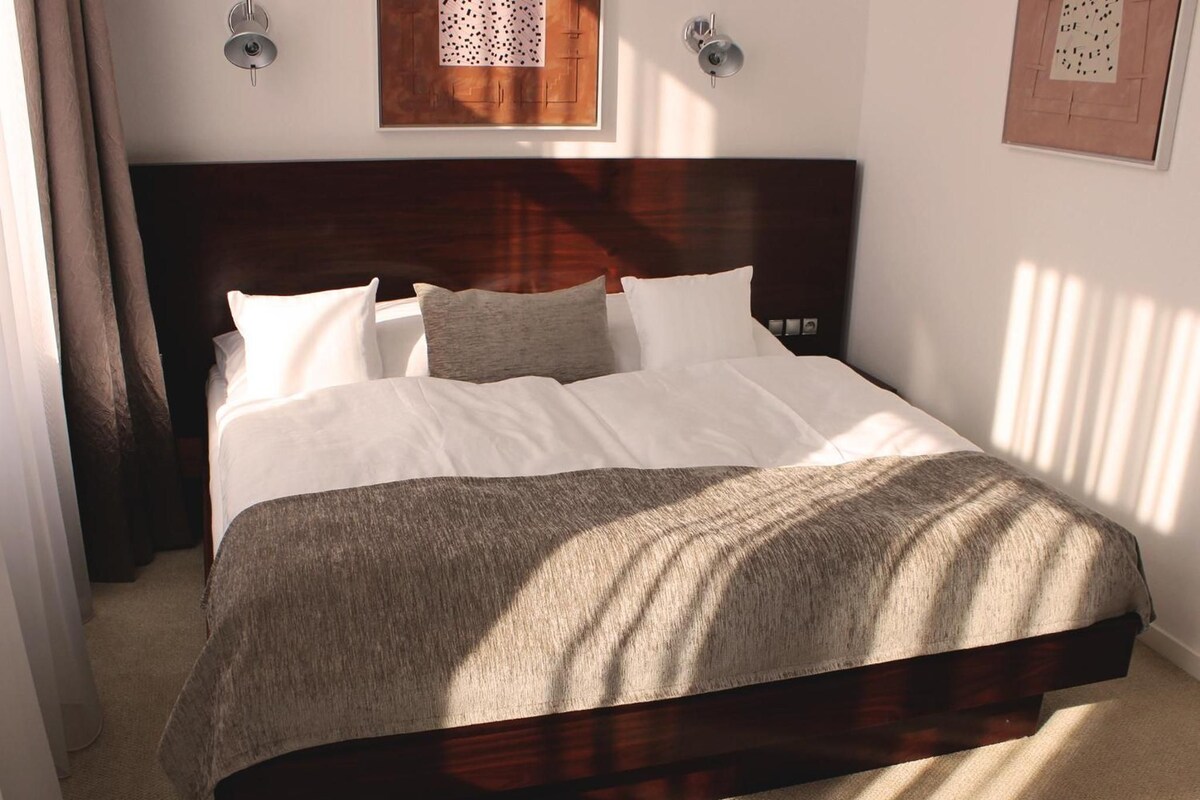 Cama bem arrumada com cabeceira escura, roupa de cama branca e manta cinza.