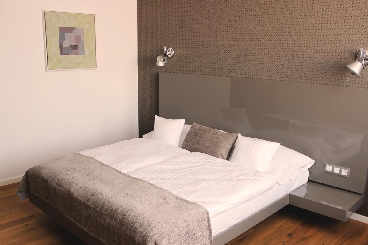 A modern bedroom with a large bed, a headboard with integrated lighting, a bedside table, and a wooden floor.