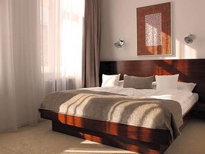 Quarto de hotel com uma cama grande, cabeceira de madeira, obra de arte emoldurada na parede e uma janela com cortinas.