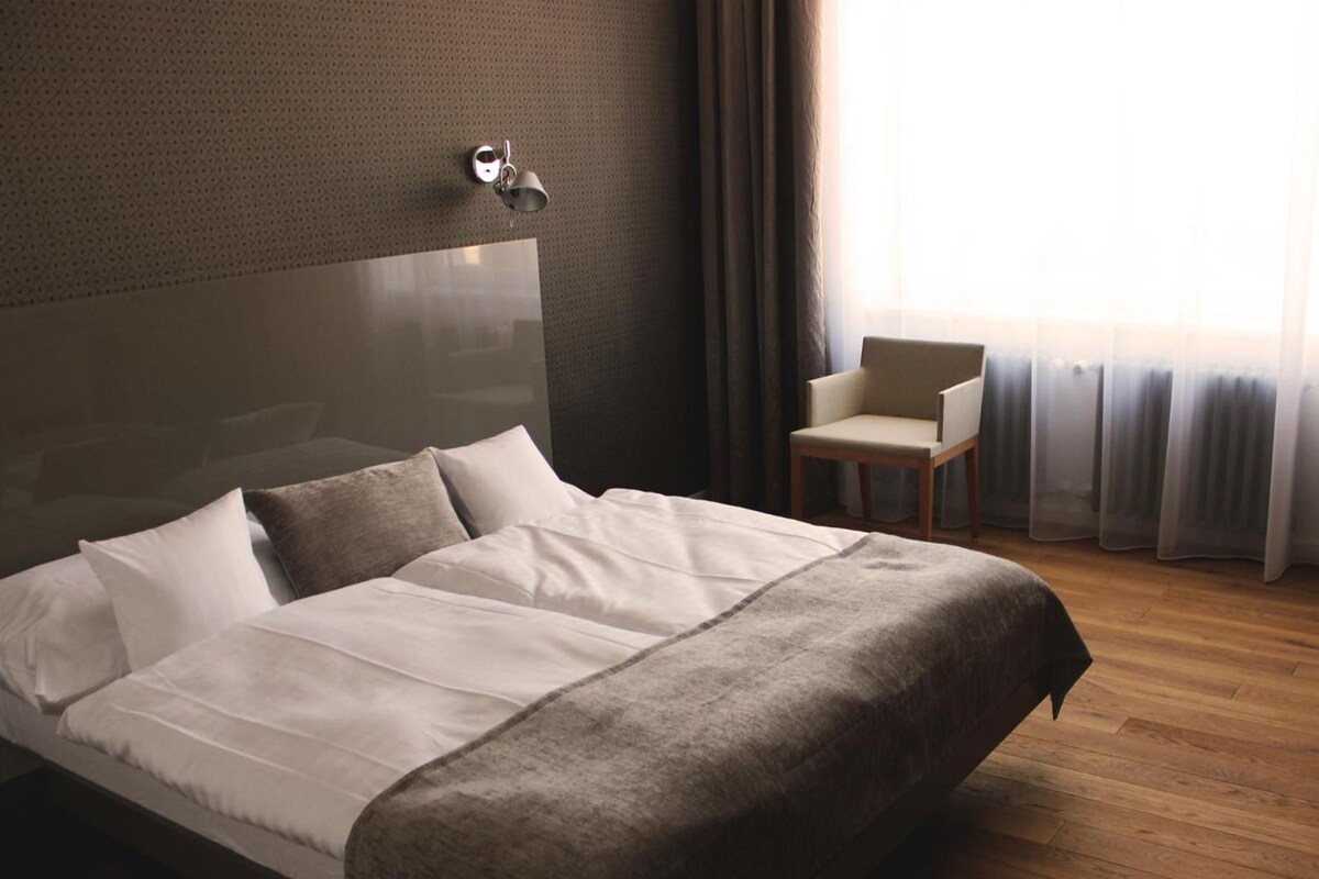 Quarto de hotel com uma cama grande, uma cadeira e uma janela com cortinas.