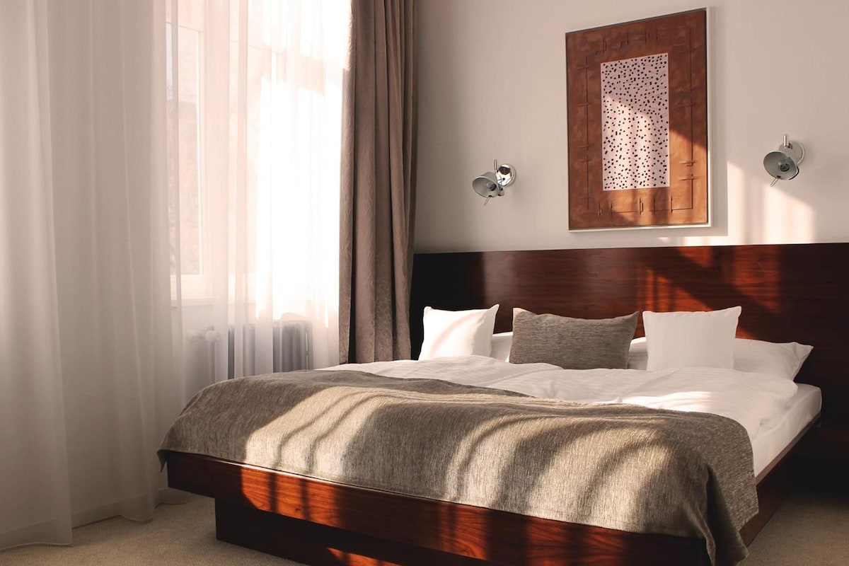A hotel room with a large bed, a wooden headboard, a window with curtains, and a wall-mounted artwork.