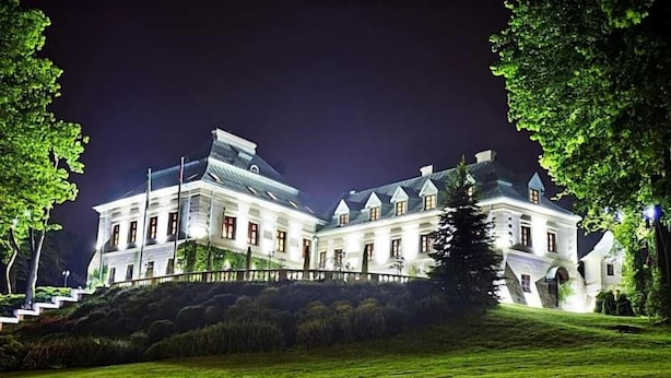 A large, illuminated building with multiple wings, surrounded by a well-maintained lawn and trees.