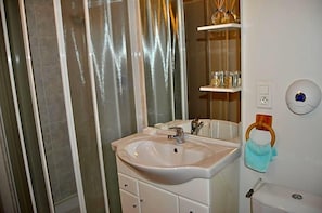 Casa de banho com lavatório branco, espelho e sanita. Há um suporte para toalhas na parede e uma prateleira com artigos de higiene pessoal.