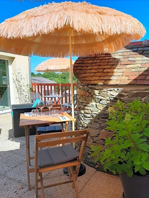 Uma área externa com mesas e cadeiras sob um toldo de palha, uma parede de pedra e um vaso de plantas.