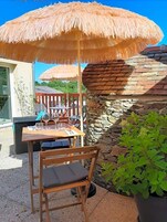 Uma área externa com mesas e cadeiras sob um toldo de palha, uma parede de pedra e um vaso de plantas.