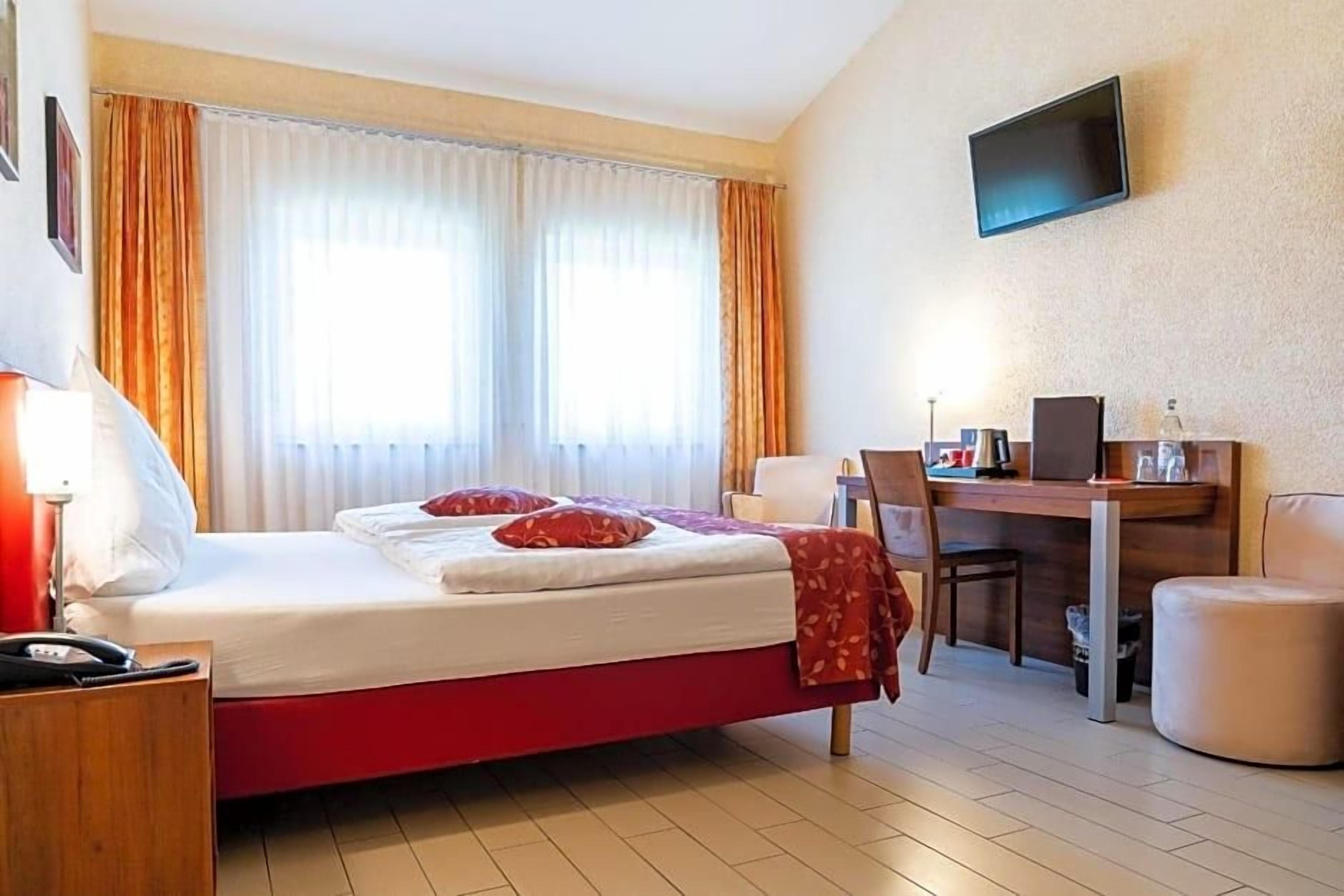 A hotel room with a bed, a desk, a chair, a TV, and a window with curtains.