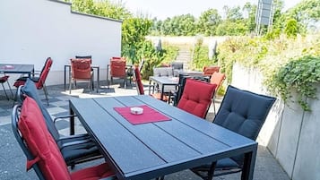Eine Terrasse im Freien mit Tischen und Stühlen, umgeben von Grünpflanzen und einer weißen Mauer.