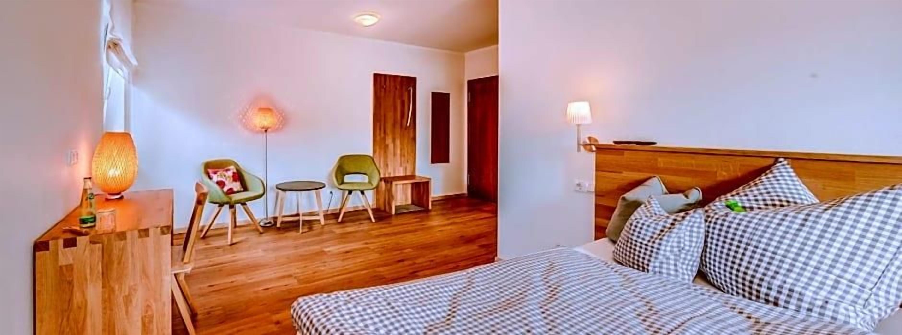 Ein Hotelzimmer mit Holzboden, einem Bett mit kariertem Bettzeug, einem Nachttisch mit Lampe, zwei Stühlen und einem kleinen Tisch.