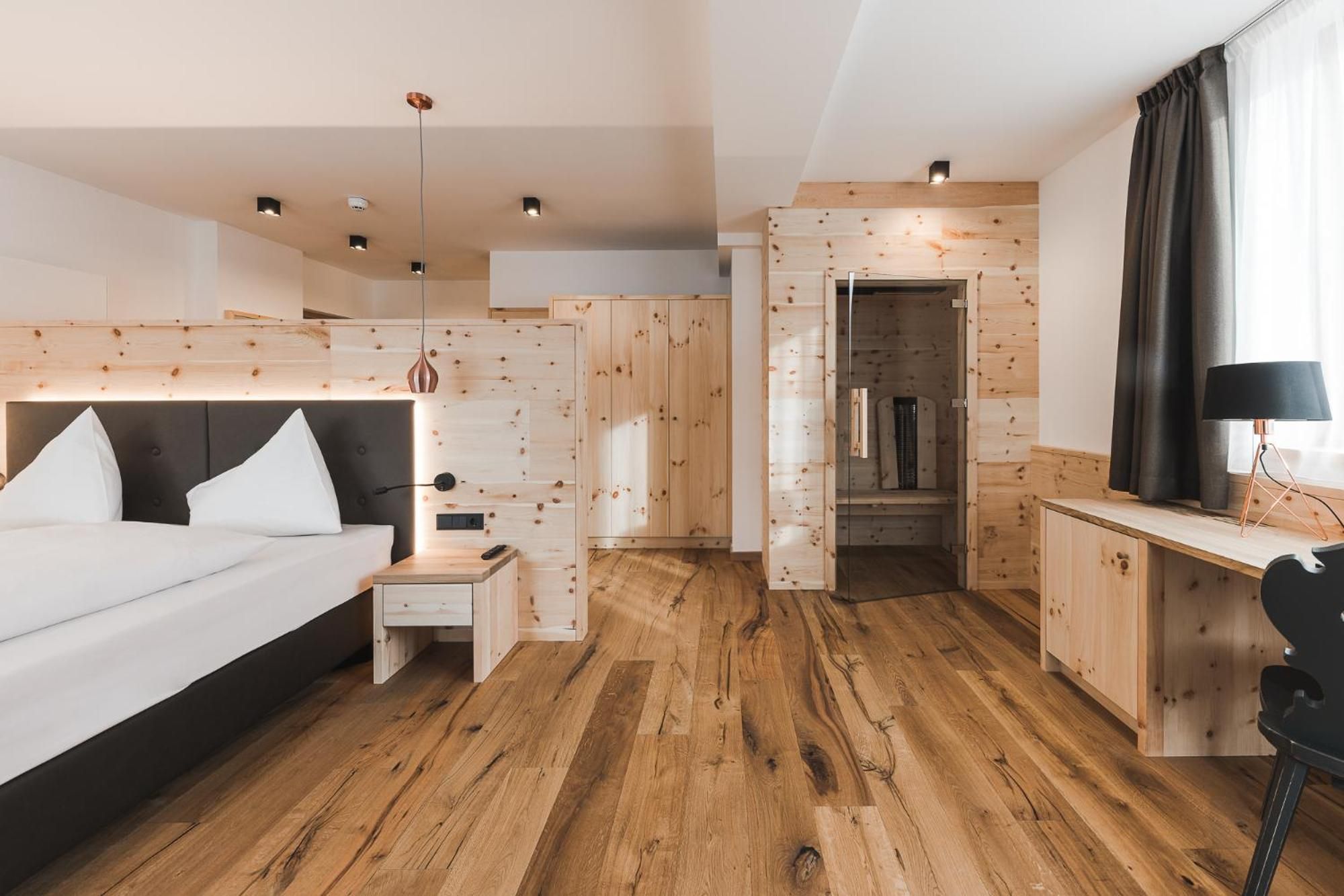 Una camera d'albergo moderna con pavimento in legno, un letto con biancheria bianca, un comodino, una scrivania con una lampada e un armadio.