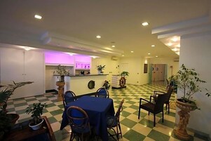 Un comedor de hotel con suelo a cuadros, una mesa azul y sillas. Se observan plantas en macetas y una cocina al fondo.