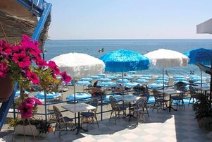 Un patio frente al mar con mesas y sillas, sombrillas azules y vistas al océano.