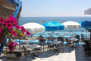 Un patio frente al mar con mesas y sillas, sombrillas azules y vistas al océano.