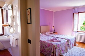 A room with a bathroom, two beds with floral bedding, a window with curtains, and a radiator.