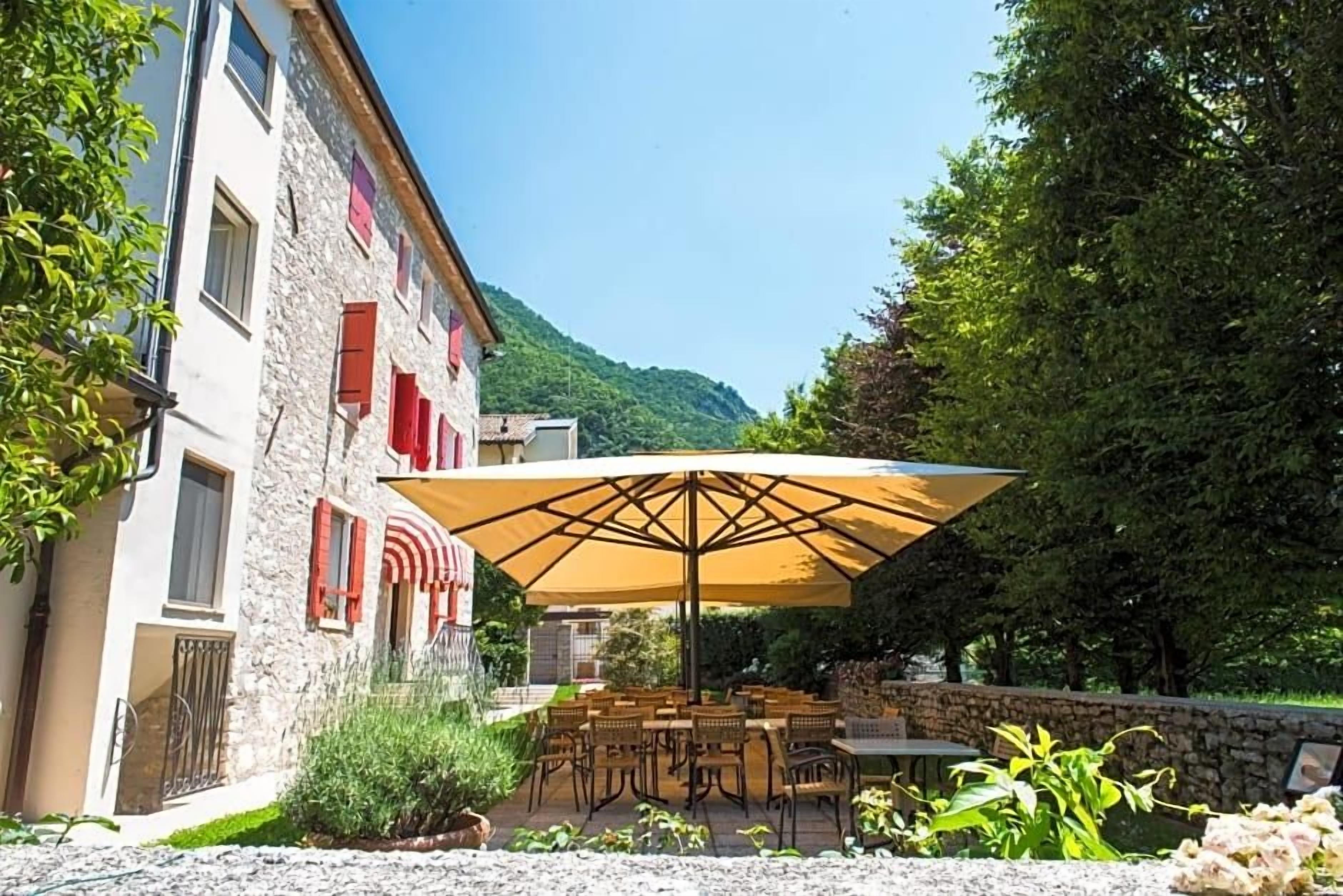 Un edificio de piedra con contraventanas rojas, un patio con mobiliario exterior y una montaña al fondo.