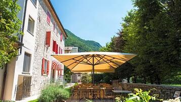 Ein Steingebäude mit roten Fensterläden, eine Terrasse mit Gartenmöbeln und ein Berg im Hintergrund.