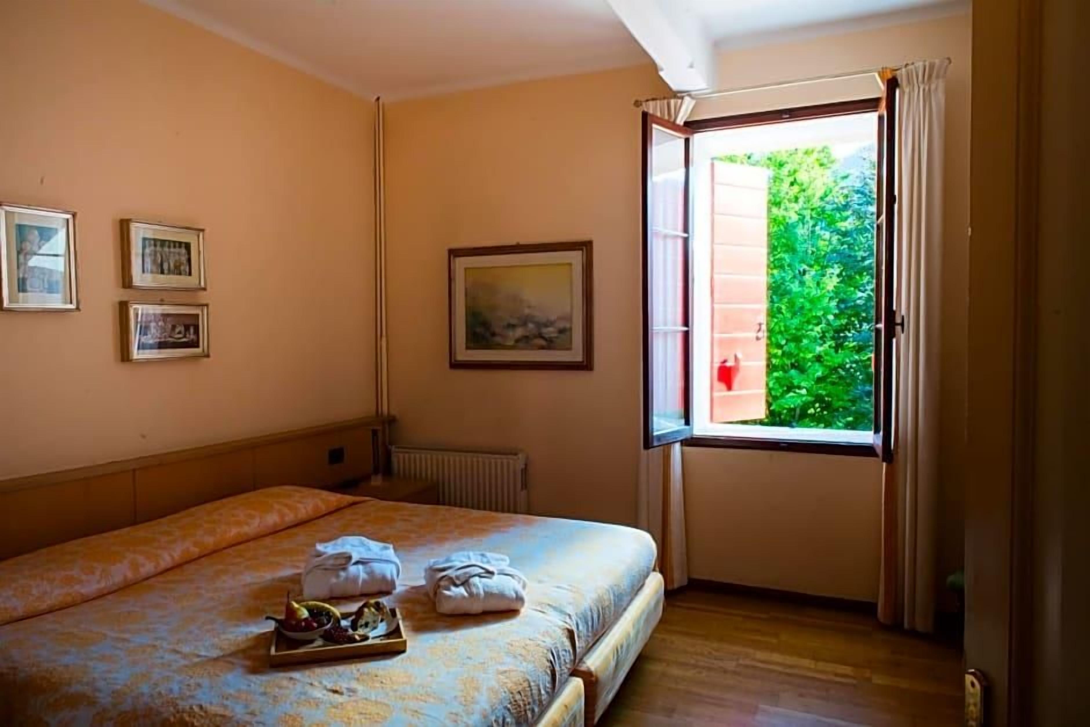 Una habitación de hotel con una cama, una ventana con cortinas, un radiador y cuadros en la pared.