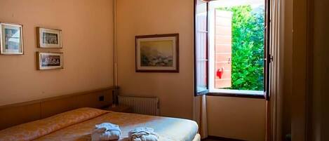 Una camera d'albergo con un letto, una finestra con tende, un termosifone e quadri appesi al muro.