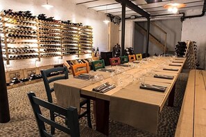 A long dining table set for a meal, surrounded by chairs, with a wine rack in the background.