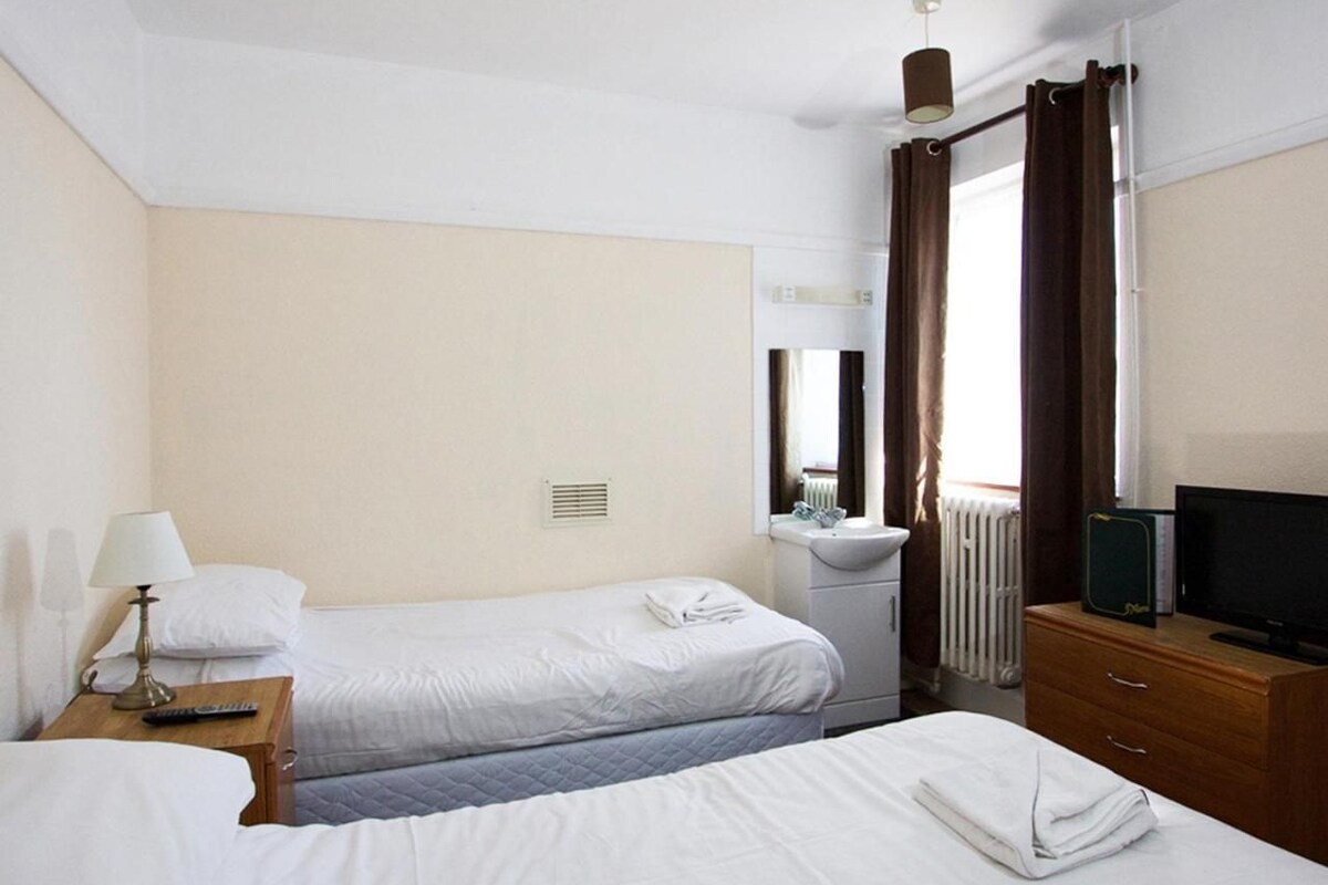 Une chambre d’hôtel avec deux lits, une table de chevet avec une lampe, une commode, un téléviseur et une fenêtre avec des rideaux.