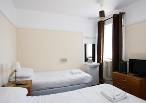 Une chambre d’hôtel avec deux lits, une table de chevet avec une lampe, une commode, un téléviseur et une fenêtre avec des rideaux.