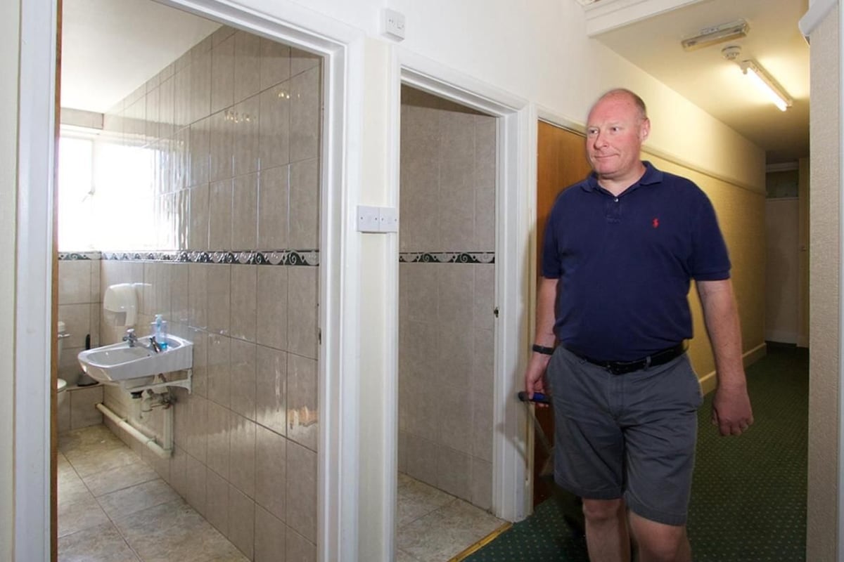 A man in a blue polo shirt and gray shorts standing in a hallway with a bathroom visible to the left.
