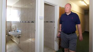 A man in a blue polo shirt and gray shorts standing in a hallway with a bathroom visible to the left.