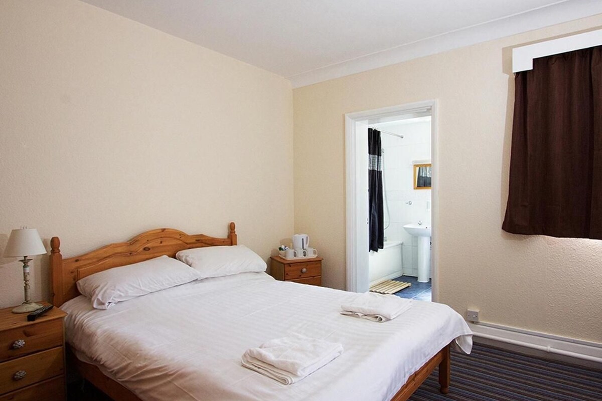 Une chambre d’hôtel avec un lit simple, une table de chevet avec une lampe et une télécommande, une salle de bain visible à travers une porte ouverte et une fenêtre avec des rideaux.