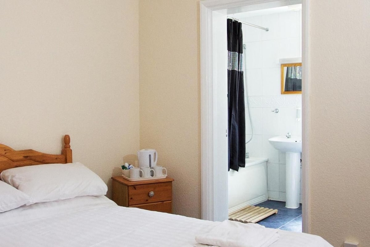 A bedroom with a wooden headboard, a bed with white linens, a nightstand with a tray of cups and a kettle, and a view into a bathroom with a sink and shower curtain.