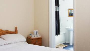 A bedroom with a wooden headboard, a bed with white linens, a nightstand with a tray of cups and a kettle, and a view into a bathroom with a sink and shower curtain.