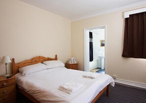 Une chambre d’hôtel avec un lit simple, une table de chevet avec une lampe et une télécommande, une salle de bain visible à travers une porte ouverte et une fenêtre avec des rideaux.