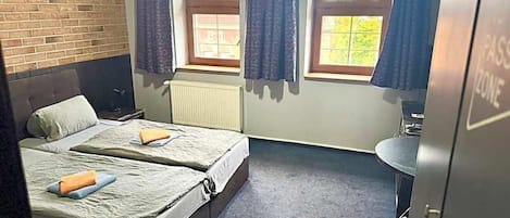 A room with two beds, a window with curtains, a radiator, a sink, and a door labeled 'PASSPORT ZONE'.