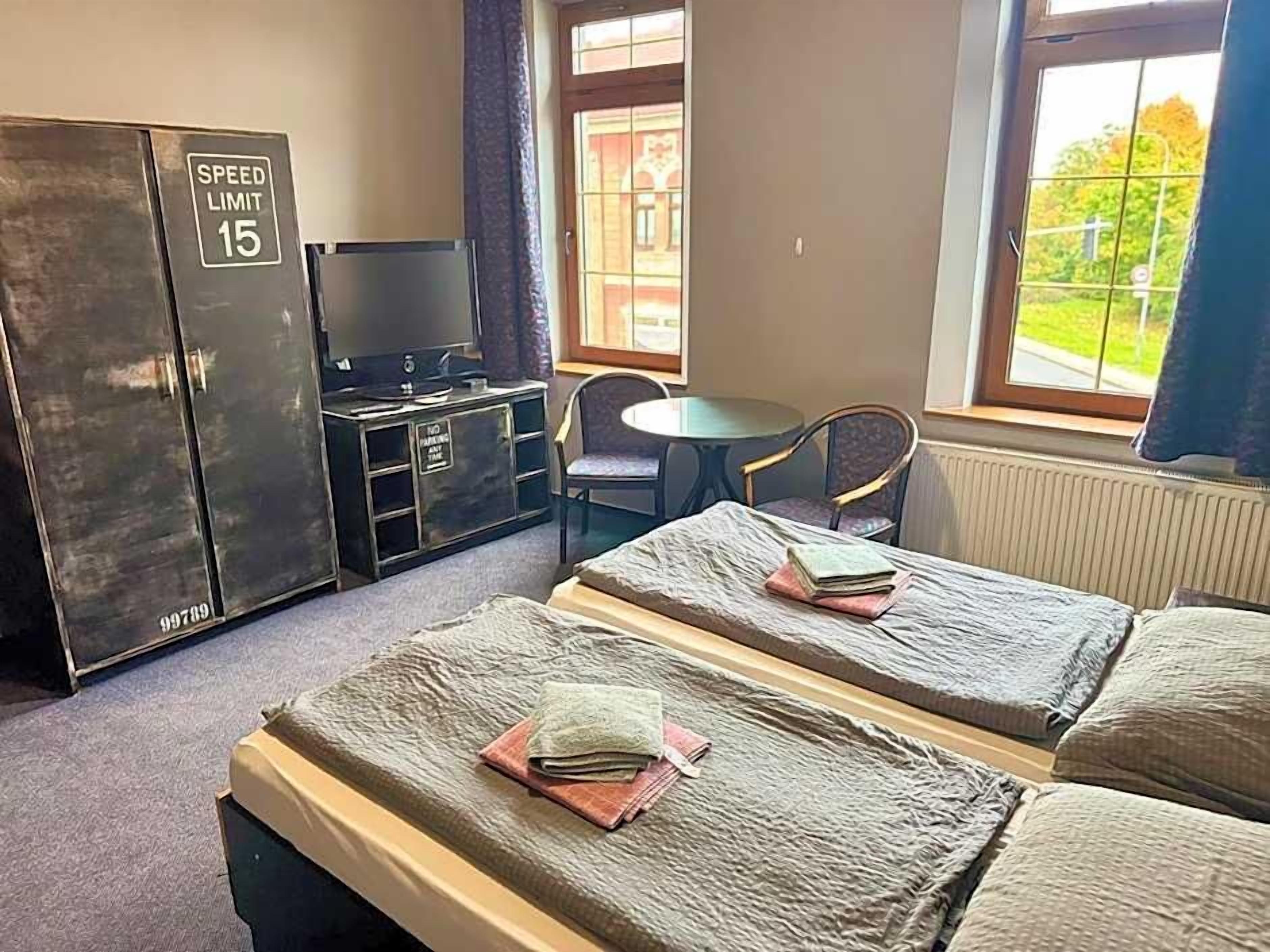 Ein Hotelzimmer mit zwei Betten, einem Schreibtisch mit Computer, einem Stuhl und einem Fenster mit Blick auf Grünflächen.