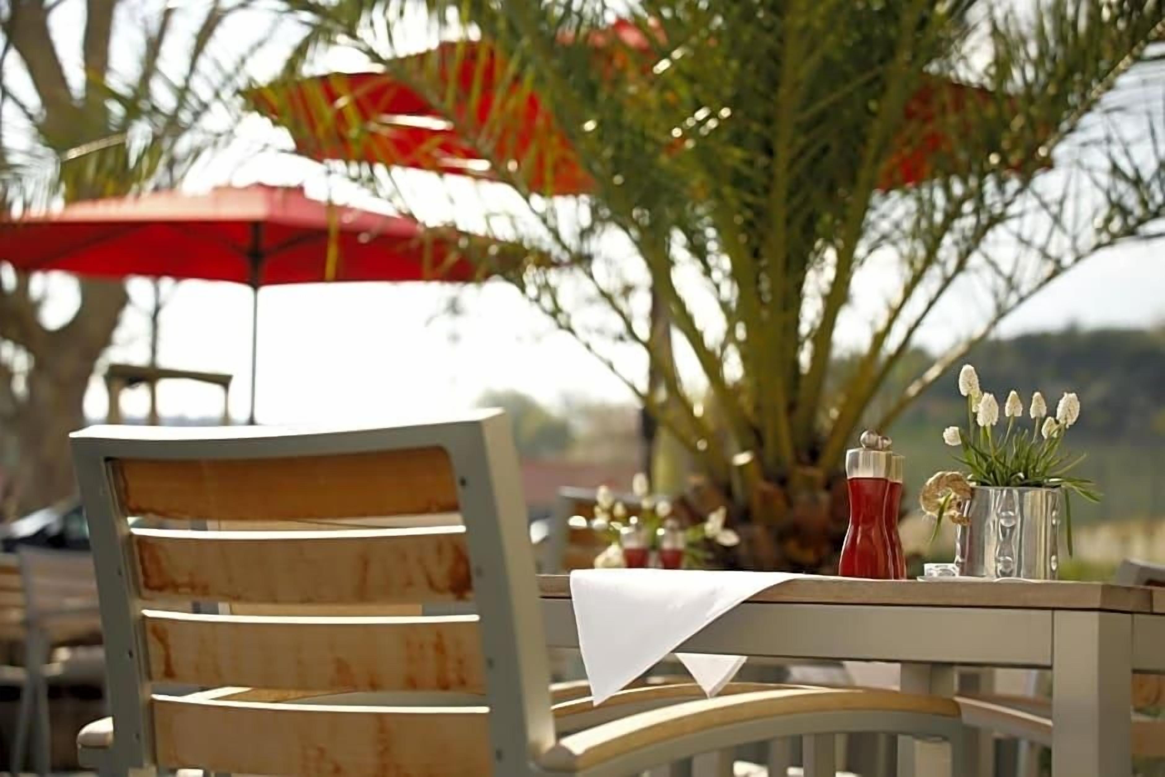 A table with a white tablecloth, a chair with a wooden backrest, and a red umbrella in the background.