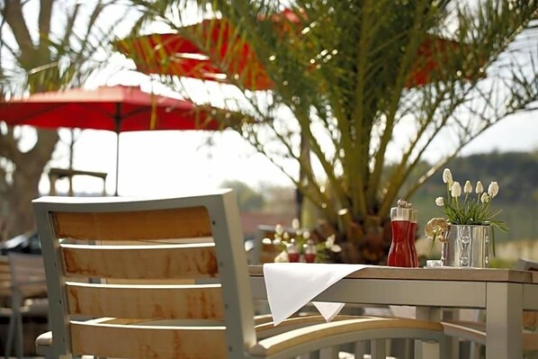 A table with a white tablecloth, a chair with a wooden backrest, and a red umbrella in the background. - Weinhaus Henninger (Kallstadt)