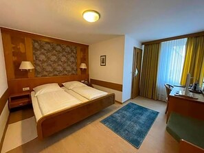 Een hotelkamer met een tweepersoonsbed, nachtlampjes, een bureau met stoel, een klein tafeltje, een raam met gordijnen en een kunstwerk aan de muur.