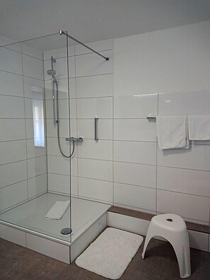 Ducha con cerramiento de vidrio, cabezal de ducha de mano, inodoro blanco y un pequeño taburete blanco.