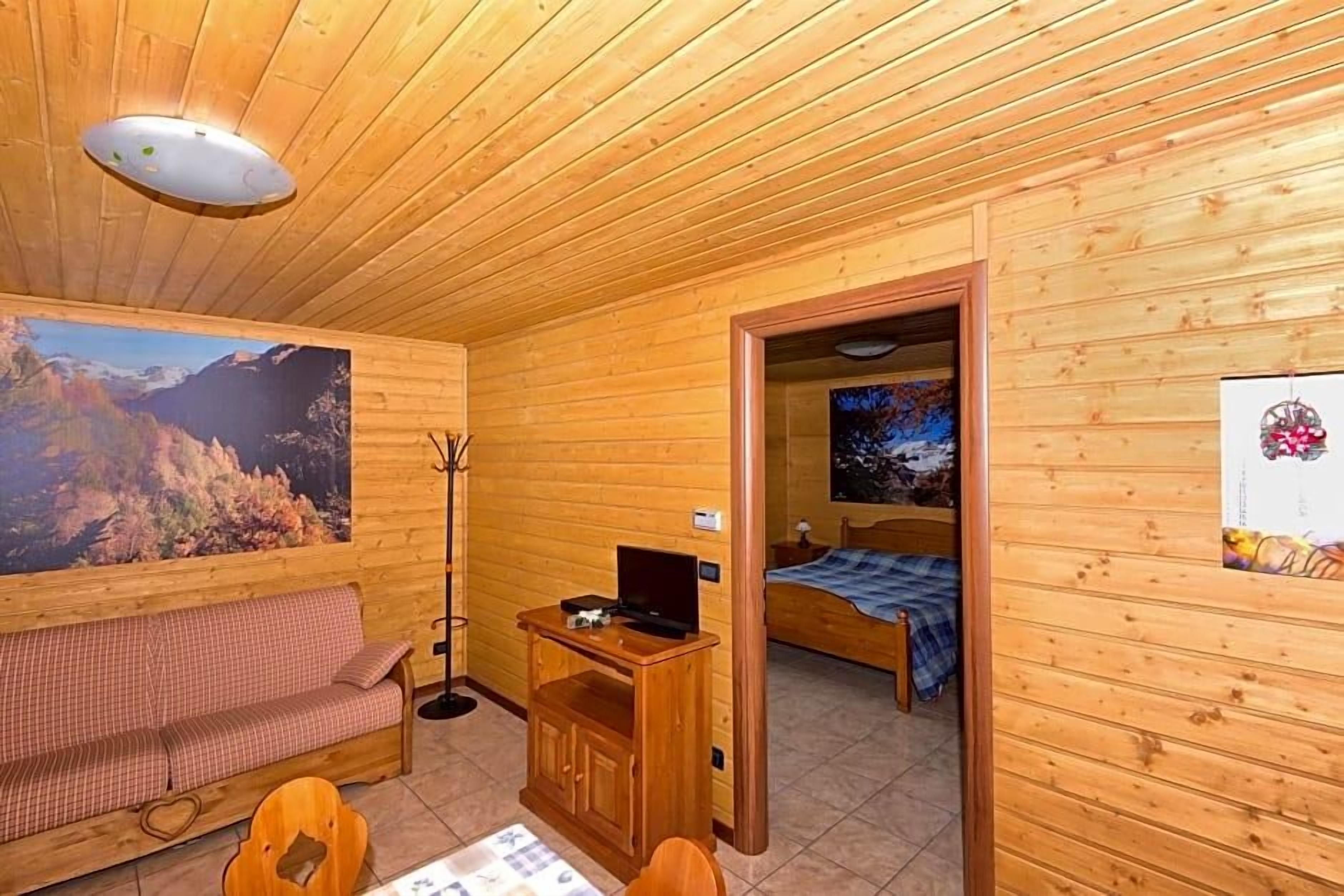 A wooden cabin interior with a sofa, a small table, a bed, and a painting of a mountain landscape.