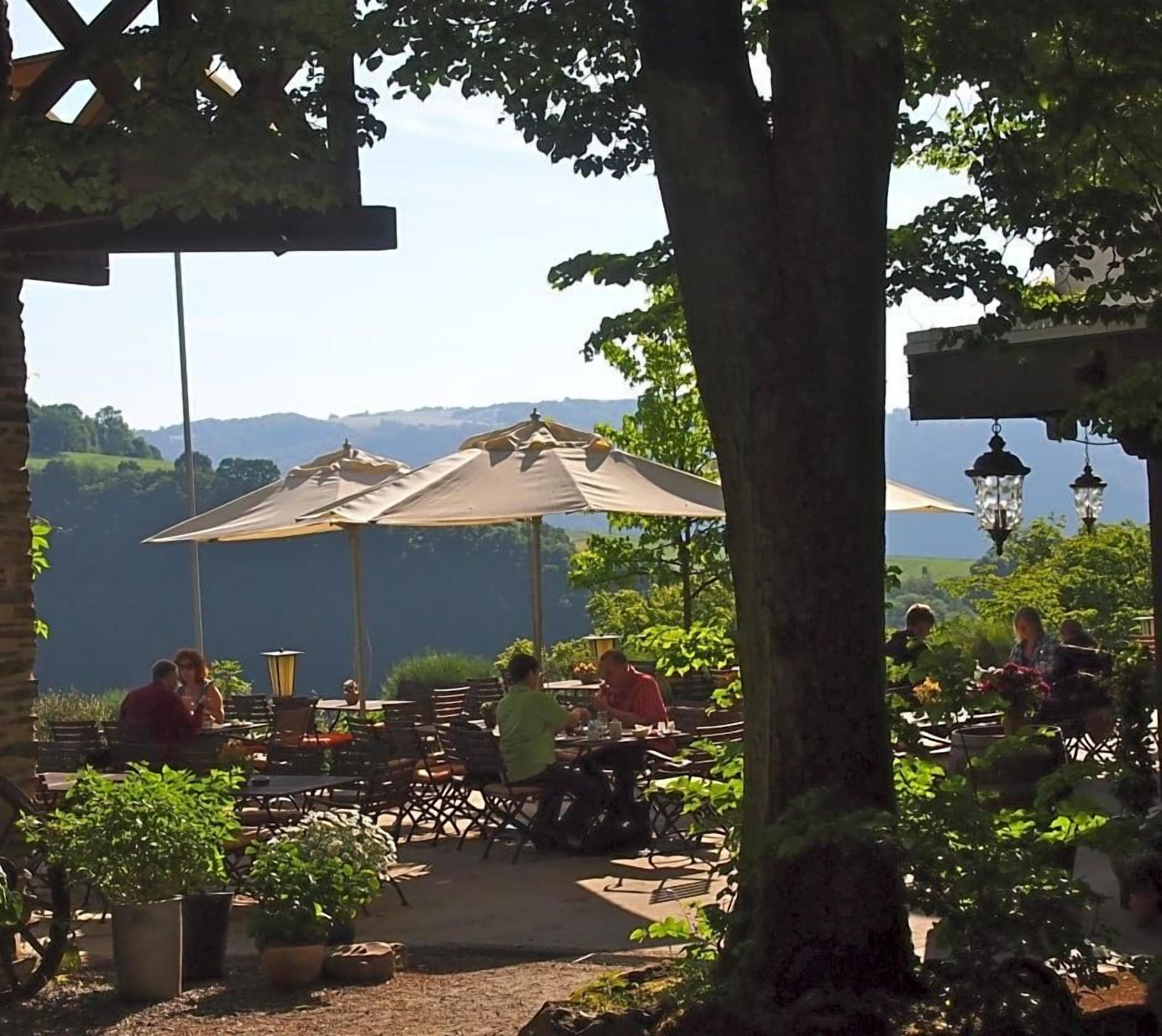 Een terras met tafels en parasols, omgeven door groen en met uitzicht op een prachtig landschap.