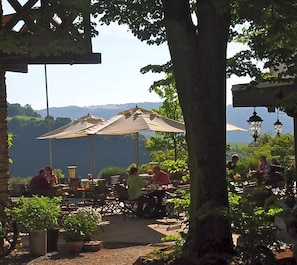 Un espacio de comedor al aire libre con mesas y sombrillas, rodeado de vegetación y vistas a un paisaje pintoresco.