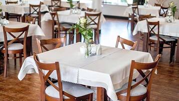 Interior de restaurante con sillas y mesas de madera, manteles blancos y un jarrón con flores sobre la mesa.