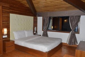 A bedroom with a wooden ceiling, a bed with white bedding, a nightstand, a chair, and a window with curtains. - The Retreat (Puri)