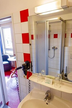 Un baño con ducha, lavabo y espejo. Paredes con azulejos rojos y blancos, y una pequeña mesa con una silla al fondo.