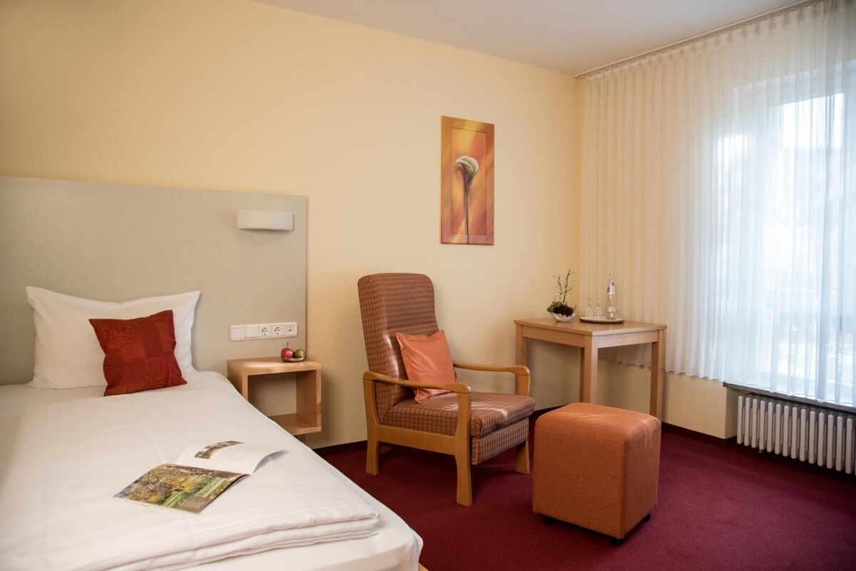 A hotel room with a bed, a chair, a small table, and a window with curtains.