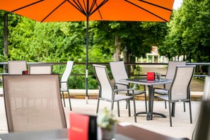 An outdoor seating area with tables and chairs, an orange umbrella, and a view of a garden with trees and a fountain. - Hotel Kurhaus Bad Bocklet (Bad Bocklet)