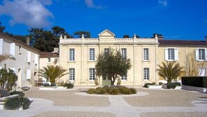 Un edificio grande de estilo clásico con fachada simétrica, entrada central y jardín formal con senderos de grava y setos podados.