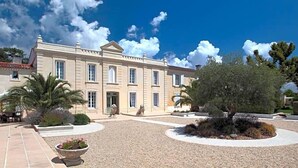 Un edificio grande de estilo clásico con fachada simétrica, una entrada central y un patio de grava con una palmera y un parterre floral.
