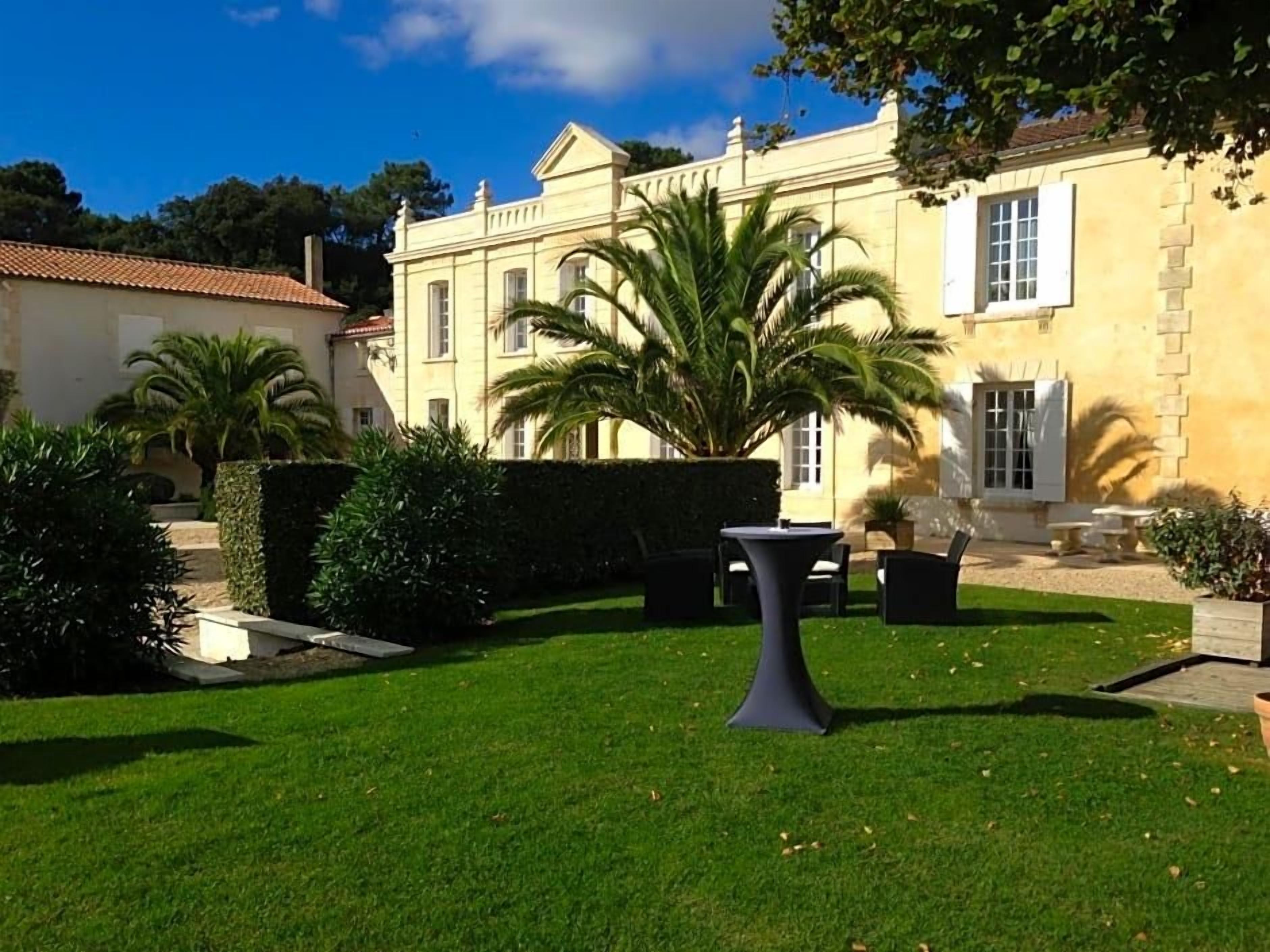 A well-maintained garden with a palm tree, a table, and chairs, and a building with a classic architectural style in the background.