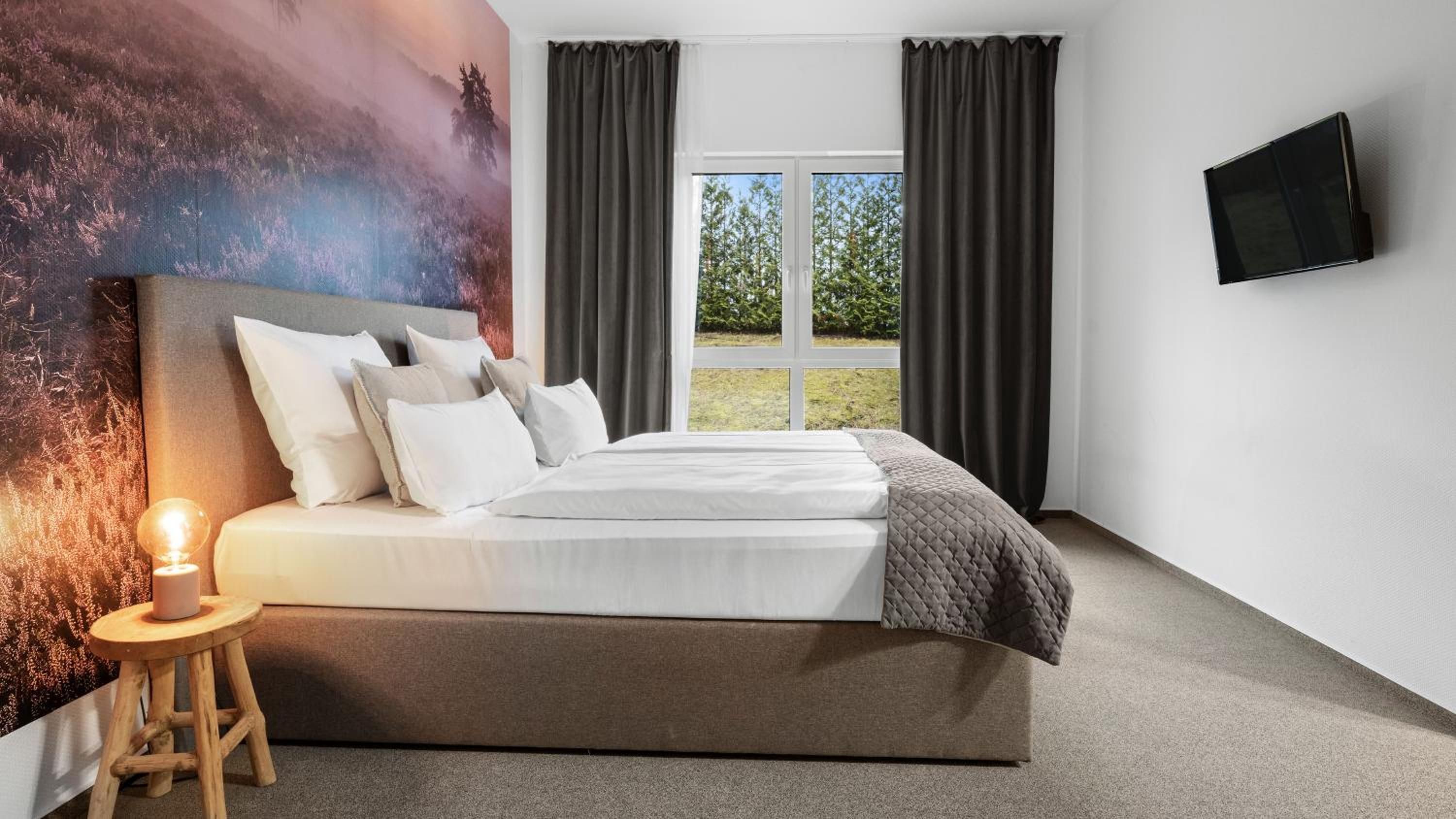 Ein modernes Schlafzimmer mit einem großen Bett, einem Nachttisch, einem an der Wand montierten Fernseher und Blick ins Freie.