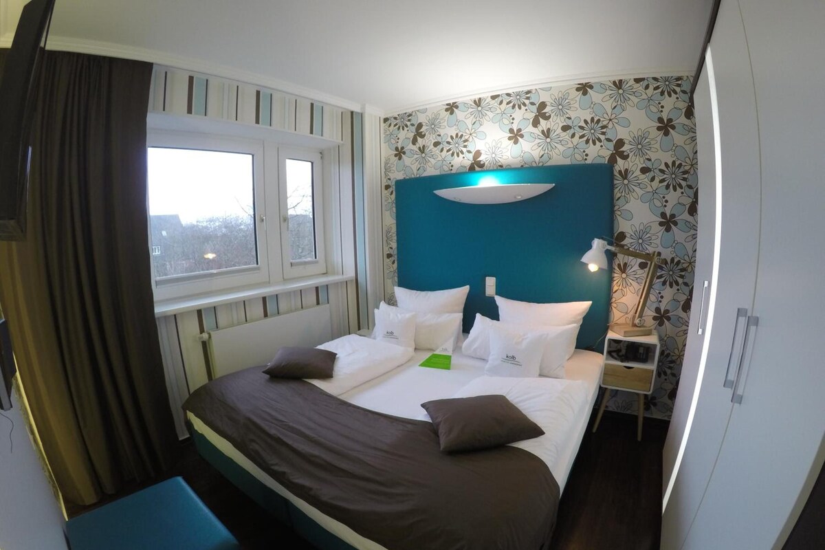 Habitación de hotel con cama, ventana, mesita de noche y pared azul.